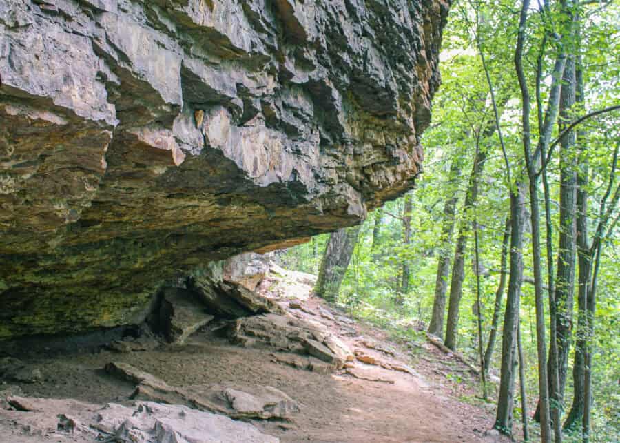 A dirt hiking trail passes beneath a large rocky overhang along a forested path in Huntsville, Alabama. The shaded rock formation creates a fun and easy spot for kids to explore along a kid friendly hiking trail.