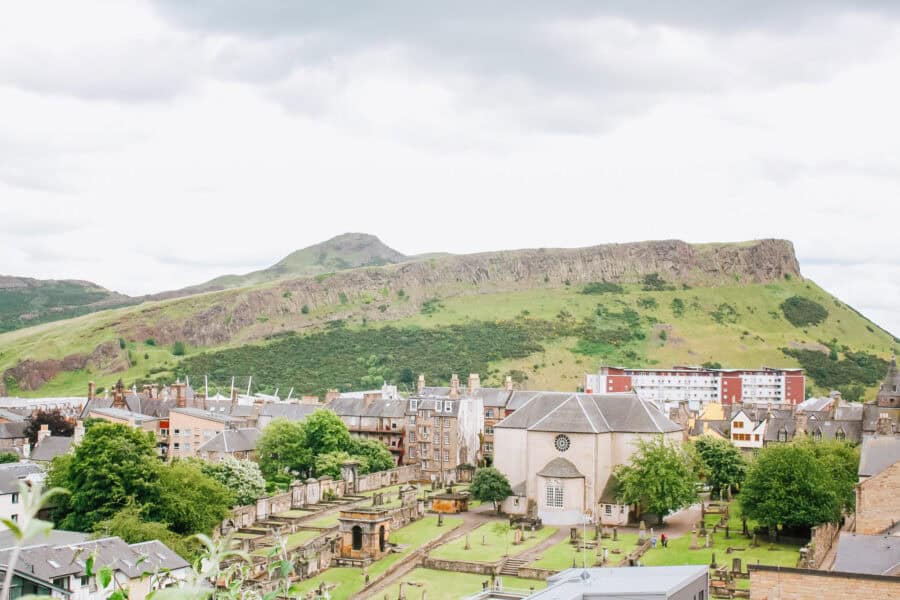 View of Arthurโs Seat, an ancient volcanic hill in Edinburgh, Scotland, rising behind the historic buildings and gravestones of a city churchyard. The lush green slopes contrast with the stone architecture and urban surroundings below.