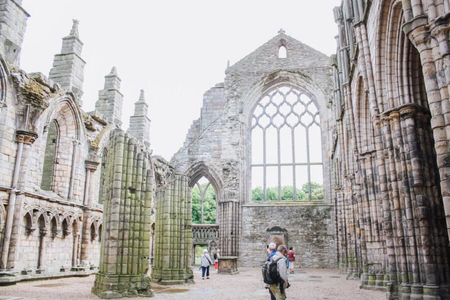 Interior ruins of Holyrood Abbey in Edinburgh, Scotland, showing weathered stone columns, arched windows, and a large gothic-style window frame open to the sky. Visitors walk through the open-air structure, surrounded by centuries-old stonework and greenery beyond the walls.