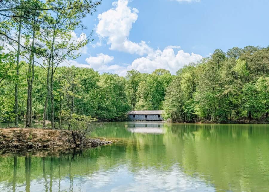 A peaceful green lake reflects tall trees and a covered wooden bridge under a bright blue sky in Huntsville, Alabama. The calm water and easy shoreline setting make it ideal for a kid friendly hiking stop.