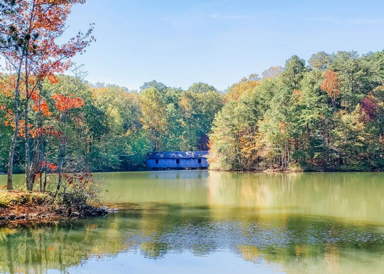A calm lake reflects colorful fall foliage with a covered wooden bridge stretching across the water, surrounded by dense forest in Huntsville, Alabama. The peaceful setting highlights a scenic, kid friendly hiking area.