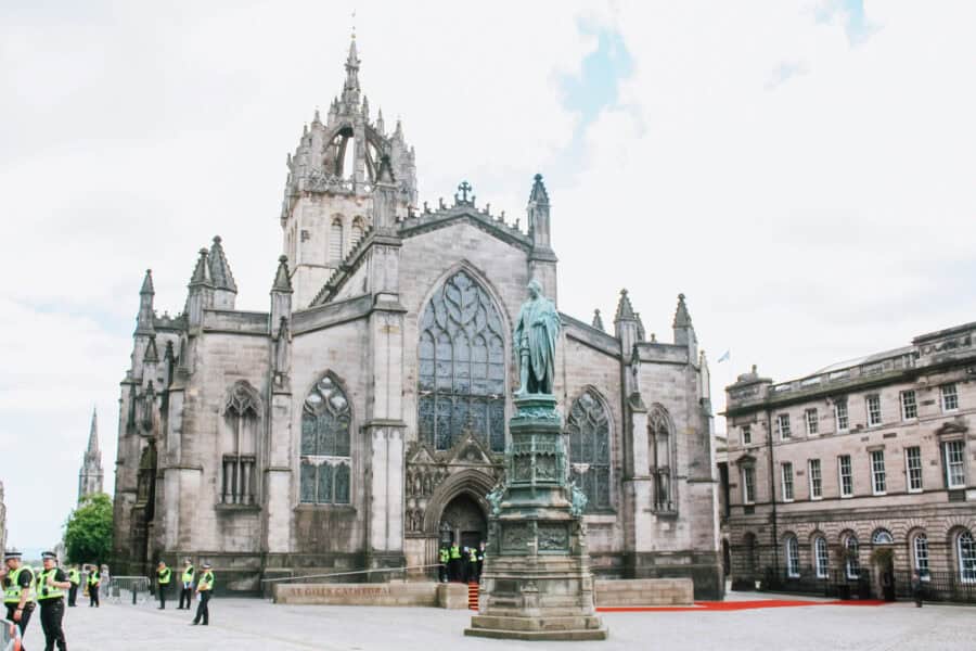 Front view of St Gilesโ Cathedral in Edinburgh, Scotland, showcasing its Gothic architecture, pointed arches, and stained glass windows. A bronze statue of Adam Smith stands in the foreground, surrounded by uniformed officers and visitors near the entrance.