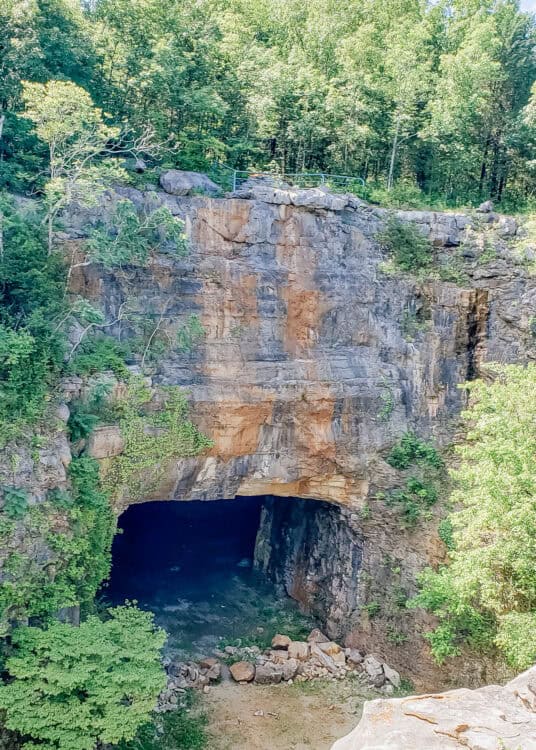 A large cave opening sits at the base of a rocky cliff surrounded by green trees along a forested trail in Huntsville, Alabama.