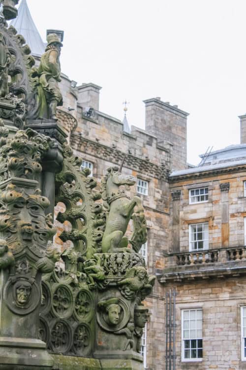 Close-up of an ornate stone fountain in the courtyard of the Palace of Holyroodhouse in Edinburgh, Scotland, featuring intricate carvings, including a rearing unicorn, human faces, and elaborate floral patterns. The palaceโs historic stone facade with tall windows and chimneys appears in the background.