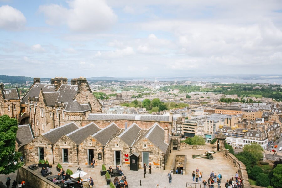 View from Edinburgh Castle overlooking the city below, with stone buildings and slate rooftops in the foreground and a panoramic cityscape stretching to the horizon. Visitors walk through the castle grounds, and a historic cannon is displayed near the edge of the fortress wall.