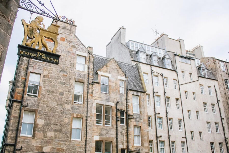 Exterior of The Writersโ Museum in Edinburgh, Scotland, with a hanging gold sign depicting a writer at a desk. The museum is set among historic stone buildings with white-framed windows and slate rooftops.