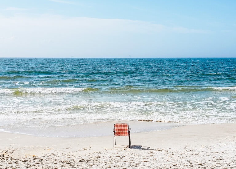 A single red beach chair sits on soft sand facing gentle ocean waves under a clear blue sky, creating a quiet and peaceful coastal scene.