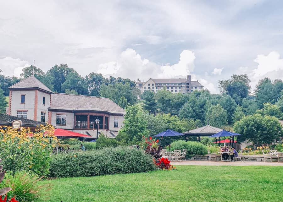 Lush gardens with colorful flowers and patio seating lead to the Biltmore Estate’s Creamery and adjacent buildings, with red and blue umbrellas providing shade for guests. In the distance, the Inn on Biltmore Estate sits atop a wooded hill under a partly cloudy sky.