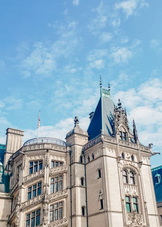 Close-up view of the ornate architecture of the Biltmore Estate in Asheville, North Carolina, showcasing intricate stone carvings, tall windows, and a steeply pitched roof topped with decorative spires. The elegant details stand out against a bright blue sky with scattered clouds.