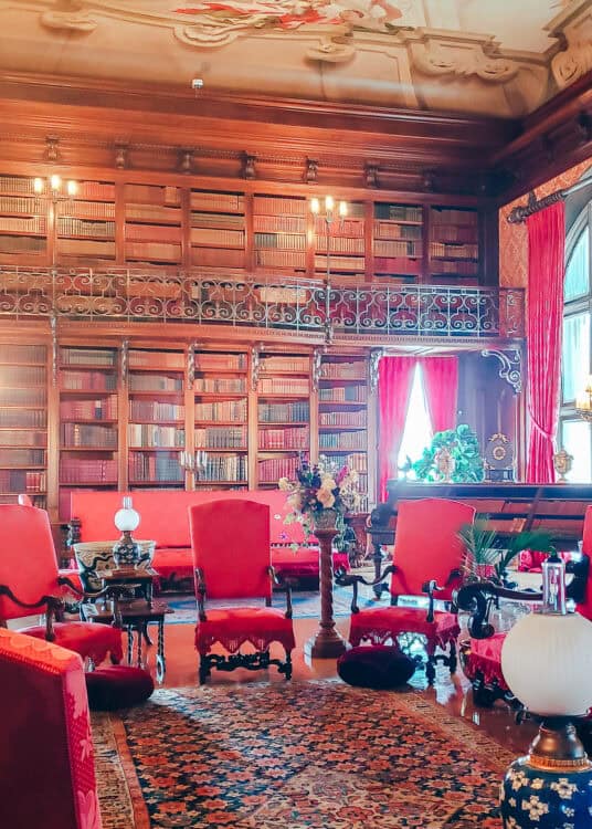 The Biltmore Estate’s opulent library features floor-to-ceiling bookshelves with a wrought iron balcony, rich red velvet chairs, ornate wood furniture, and a grand ceiling mural. Sunlight filters through tall windows draped in heavy red curtains, highlighting the room’s luxurious Gilded Age decor.