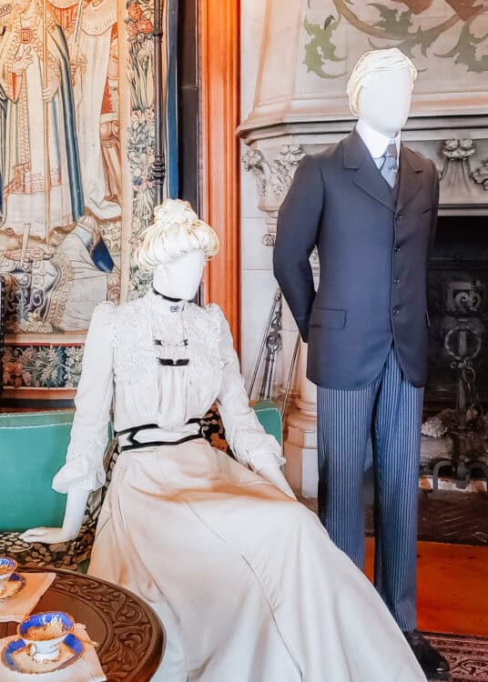 Mannequins dressed in Edwardian-era clothing are displayed in a furnished room at the Biltmore Estate, with the woman in a high-neck white lace dress and the man in a dark suit with striped trousers. They are set beside a carved fireplace and medieval-style tapestry, evoking the estate’s Gilded Age elegance.