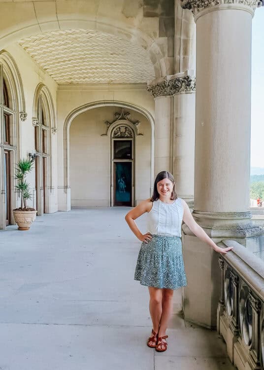 A woman poses on the grand outdoor loggia of the Biltmore Estate in Asheville, North Carolina, wearing a white sleeveless top and floral skirt. The arched windows, tall columns, and ornate ceiling details highlight the mansion's architectural elegance, with mountain views visible in the background.