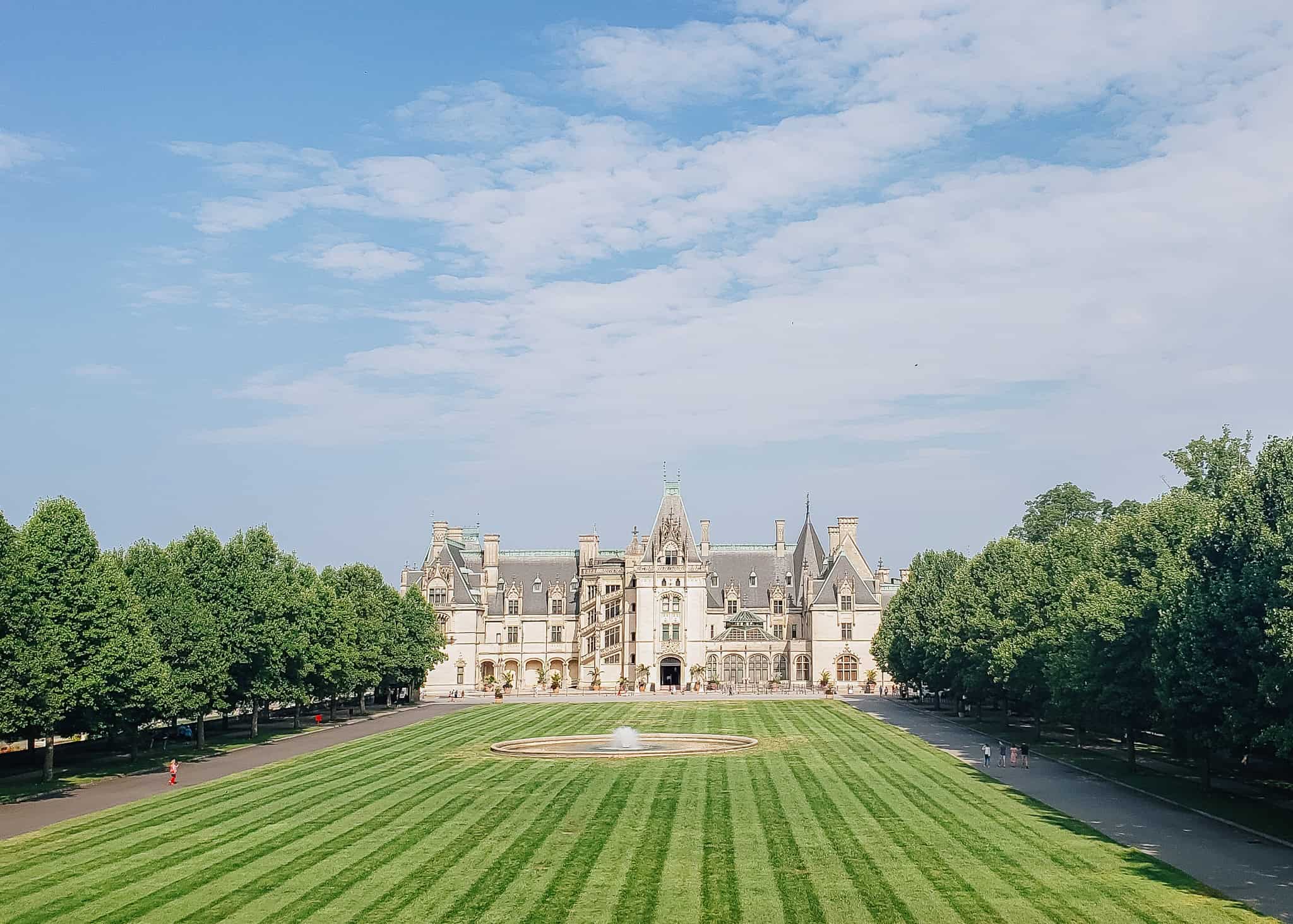 The Biltmore Estate in Asheville, North Carolina, viewed from the front lawn with manicured green grass, a round fountain, and rows of trees lining the path toward the grand château-style mansion. The sky is lightly clouded, highlighting the estate’s elegant architecture and expansive grounds.