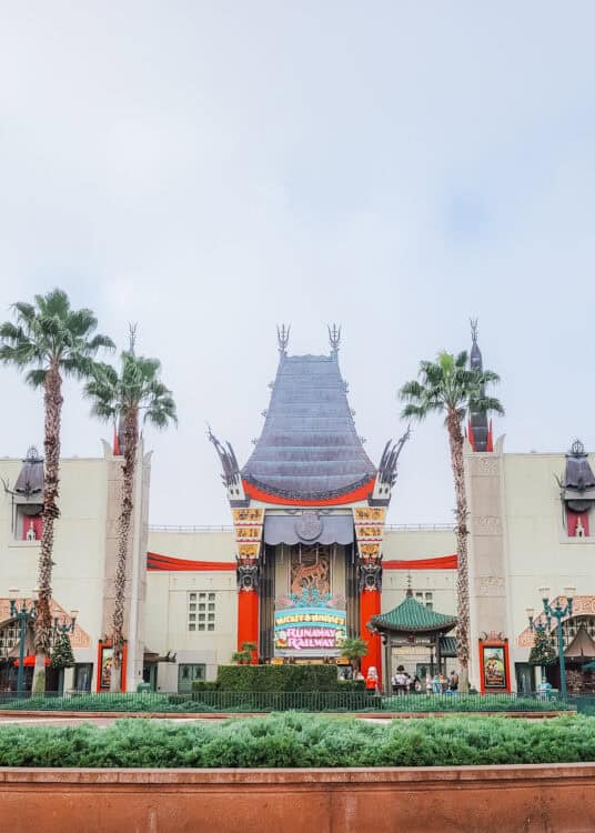 Front view of the Chinese Theatre replica at Disney's Hollywood Studios, home to Mickey & Minnie's Runaway Railway. The ornate building features traditional pagoda-style roofing, vibrant red columns, and dragon sculptures above the entrance. Palm trees frame the structure, and guests are gathered near the colorful neon marquee announcing the attraction.