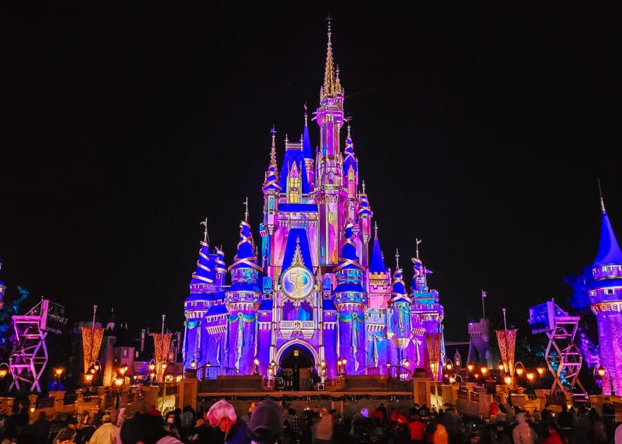 Cinderella Castle at Magic Kingdom illuminated in vibrant purple, pink, and gold during a nighttime celebration, with a large “50” emblem displayed for Walt Disney World's 50th anniversary. The crowd below watches as the castle glows against the dark sky, surrounded by festive lights and colorful turrets.