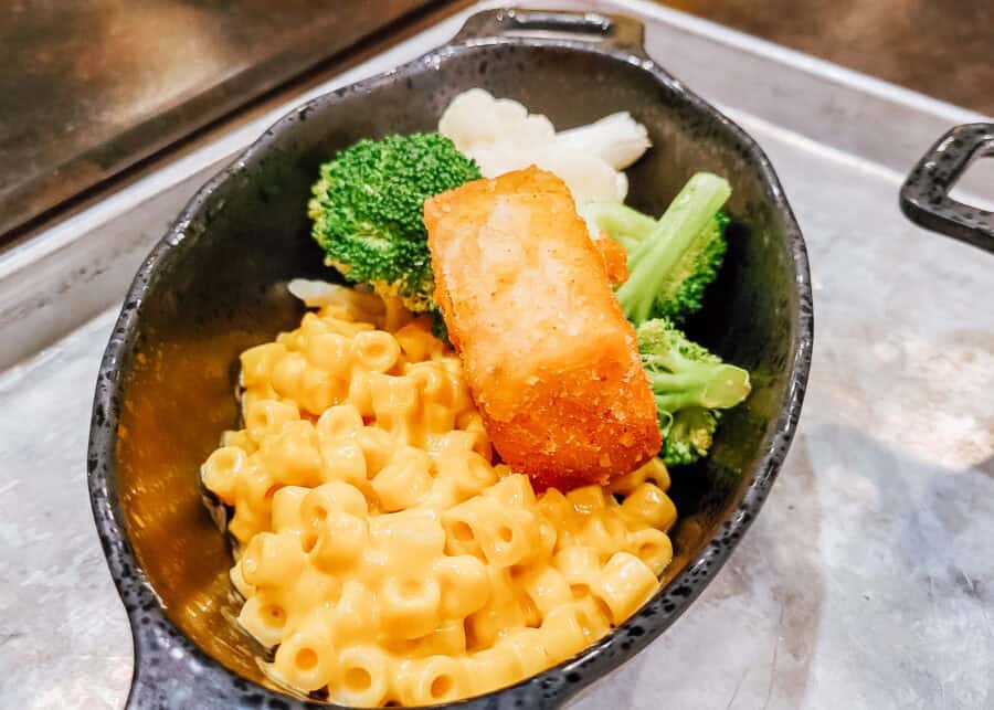 Close-up of a kid-friendly meal served in a black dish, featuring creamy macaroni and cheese, steamed broccoli and cauliflower, and a golden-brown breaded chicken nugget. The dish sits on a metal tray, with the vibrant colors and textures of the food creating an appetizing presentation.