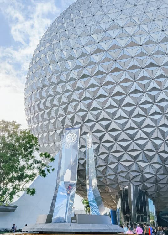 Close-up view of the iconic geodesic sphere at EPCOT in Walt Disney World, known as Spaceship Earth, with its triangular panel design glinting in the sunlight. In the foreground are reflective glass pylons featuring the EPCOT logo, and visitors are seen entering beneath the structure. The image captures a blend of futuristic architecture and theme park atmosphere.
