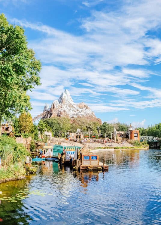 Scenic view of Expedition Everest at Disney’s Animal Kingdom, with the snow-capped mountain coaster rising in the background under a bright blue sky. In the foreground, a peaceful lagoon reflects the surrounding trees, vibrant boats, and waterfront structures, capturing the park’s immersive Himalayan-inspired setting.