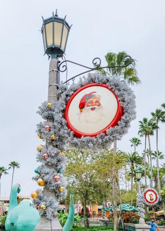 Festive holiday decoration at Disney's Hollywood Studios featuring a vintage-style Santa Claus illustration framed in red and surrounded by silver garland and colorful ornaments. The garland wraps around a tall lamppost, with palm trees and a green dinosaur statue in the background, adding a tropical twist to the Christmas display.