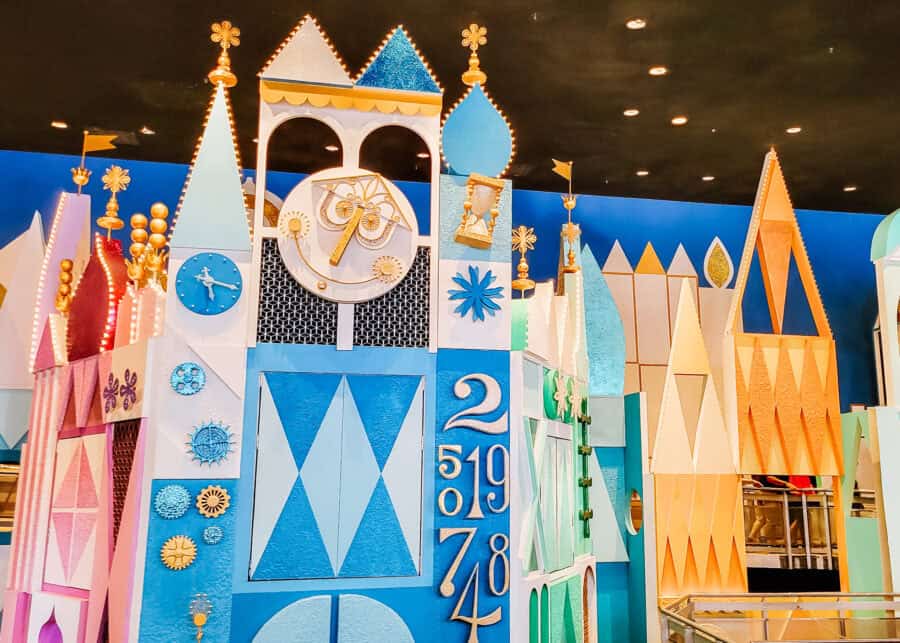 Colorful façade of the "it's a small world" ride at Magic Kingdom, featuring whimsical geometric towers, a decorative clock face with gold accents, and a playful mix of pastel blues, pinks, and gold. The stylized numbers and clockwork elements enhance the storybook charm of this beloved attraction’s iconic entrance.