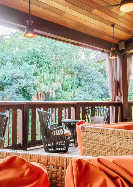 Covered outdoor lounge area with wicker and rattan seating, orange cushions, and hanging pendant lights, overlooking a lush tropical landscape filled with palm trees and dense greenery. The wooden ceiling and railing add to the relaxed, resort-style ambiance of the space.