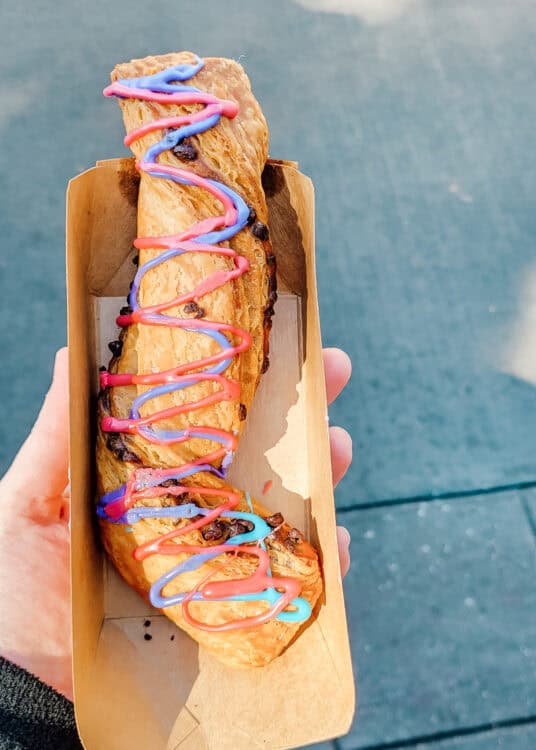 A hand holds a twisted pastry in a cardboard tray, topped with colorful drizzles of pink, purple, and blue icing. The golden-brown pastry appears flaky and studded with chocolate chips, presented outdoors in natural sunlight.