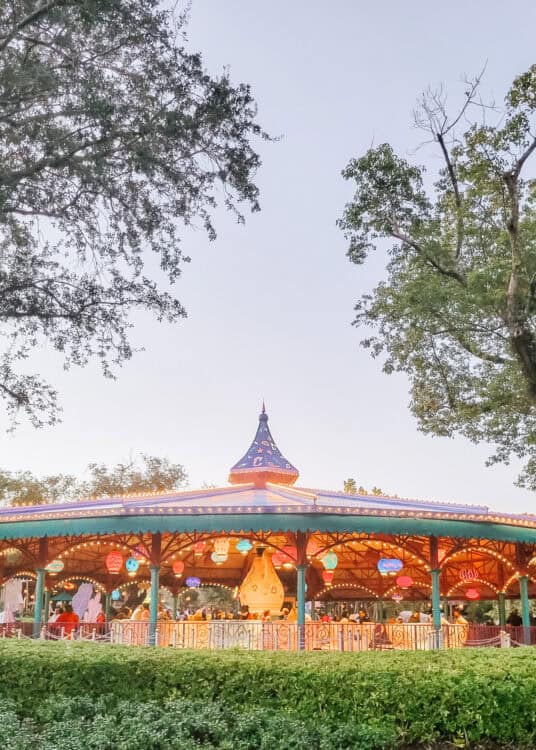 A colorful, illuminated outdoor ride at dusk featuring spinning teacups under a whimsical pavilion roof decorated with hanging lanterns. Surrounded by trees and greenery, the scene captures the Mad Tea Party attraction from Alice in Wonderland at a Disney theme park.