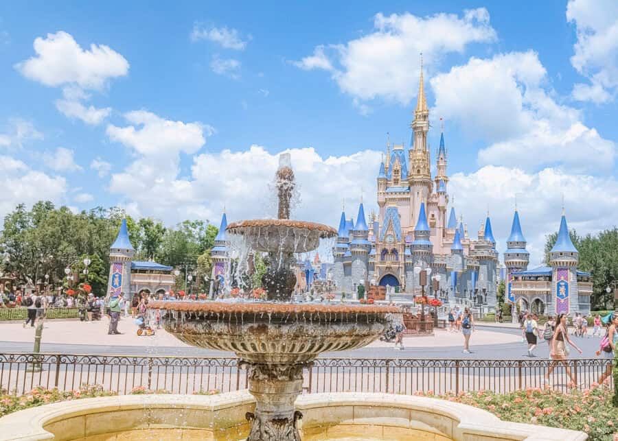 A picturesque view of Cinderella Castle at Walt Disney World's Magic Kingdom, framed by a decorative water fountain in the foreground. The sky is bright blue with scattered clouds, and guests are walking around the courtyard surrounded by colorful flower beds and medieval-style towers with blue spires.
