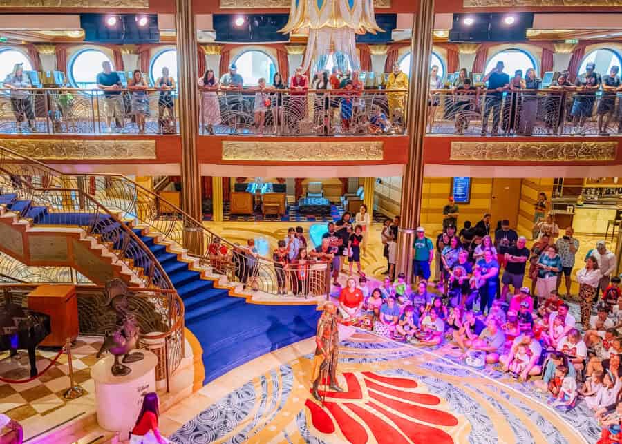Vibrant scene inside the grand atrium of a Disney cruise ship, where a costumed performer entertains a seated crowd of children and adults. The space features elegant gold accents, a sweeping staircase with royal blue carpet, ornate railings, and a large chandelier. Onlookers also watch from an upper-level balcony with large round windows.