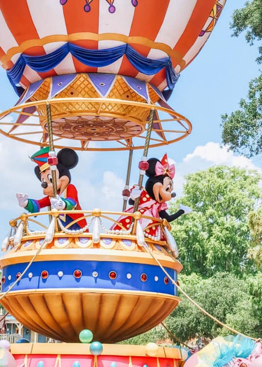 Mickey Mouse and Minnie Mouse wave from a colorful hot air balloon float during a parade at Magic Kingdom, surrounded by bright circus-style decorations and trees under a sunny blue sky.