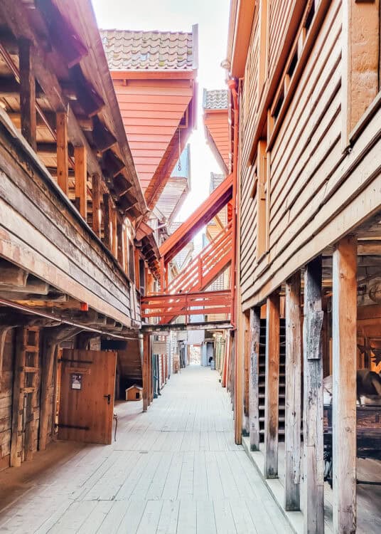 Narrow wooden alleyway between historic buildings at Bryggen in Bergen, Norway, with weathered timber walls, exposed beams, and red-painted staircases connecting the upper levels. A wooden plank walkway runs through the quiet passage beneath overhanging structures and tiled roofs.