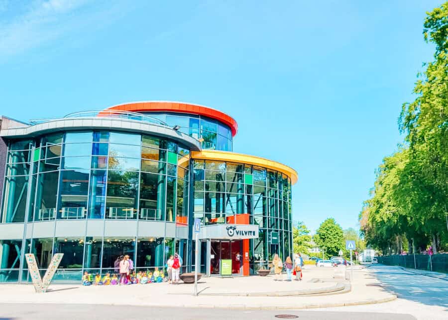 A modern circular glass building with curved walls and colorful trim in orange and yellow stands along a sunny street. A group of children wearing bright vests sit outside near the entrance, while a few adults stand nearby. Trees line the sidewalk under a clear blue sky.