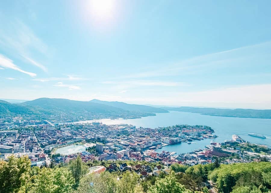 Panoramic view of a coastal city with colorful buildings clustered around a wide harbor and winding waterways. Green hills and mountains surround the shoreline, and boats dot the sparkling blue water under a clear sky. The photo is taken from an elevated viewpoint overlooking the city and fjord.