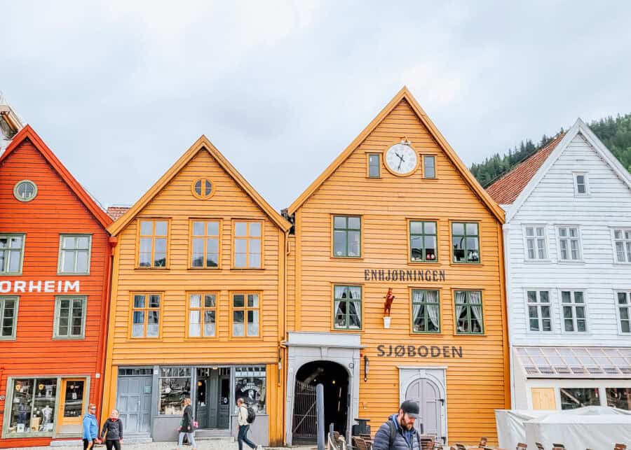Row of colorful wooden buildings with steep gabled roofs along a historic waterfront. The bright yellow facade reads ENHJรRNINGEN and SJรBODEN, with a clock centered near the roof peak and green trimmed windows. A few people walk past the shops at street level beneath the overcast sky.