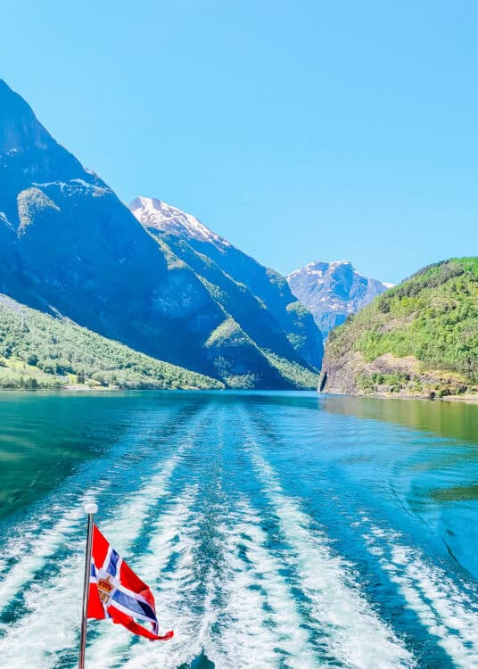 View from the back of a boat cruising through a Norwegian fjord, with the red Norwegian flag waving in the foreground. Deep blue water stretches between steep green mountains and snow capped peaks under a clear sky. The boat wake creates white ripples trailing through the calm water.