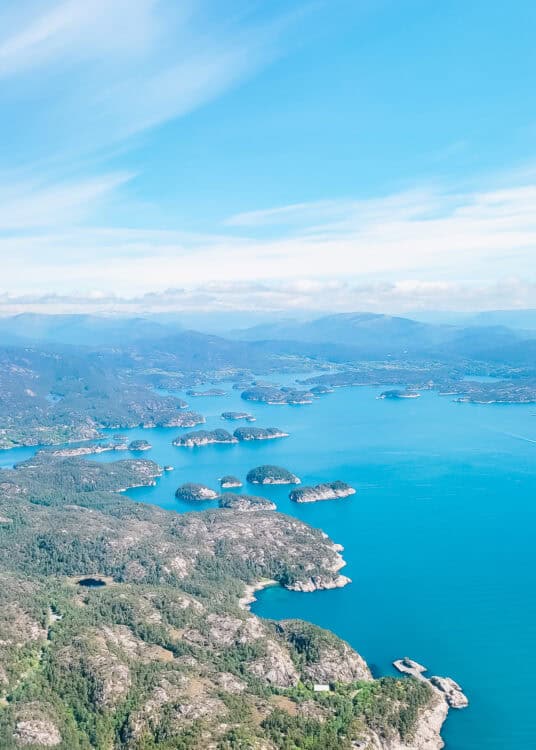 An expansive aerial view of a rugged coastline with bright turquoise water dotted by small rocky islands. Forested hills and scattered inlets stretch into the distance beneath a pale blue sky with thin, wispy clouds, creating a serene Nordic seascape.