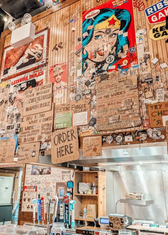 Eclectic interior of a burger restaurant covered floor to ceiling with handwritten notes stickers signs and graffiti. A large Wonder Woman artwork hangs above cardboard menus for burgers fries shakes and a sign reading "ORDER HERE".