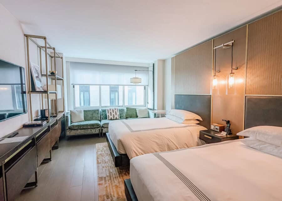 A spacious guest room at Thompson Central Park New York with two beds dressed in white linens. A green velvet sofa sits beneath a large window across from a wall mounted television and long dark console.