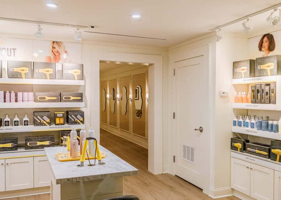 Drybar salon retail area at Thompson Central Park with shelves of Buttercup hair dryers brushes and styling products surrounding a marble display table. A hallway lined with wavy mirrors leads farther into the bright salon.