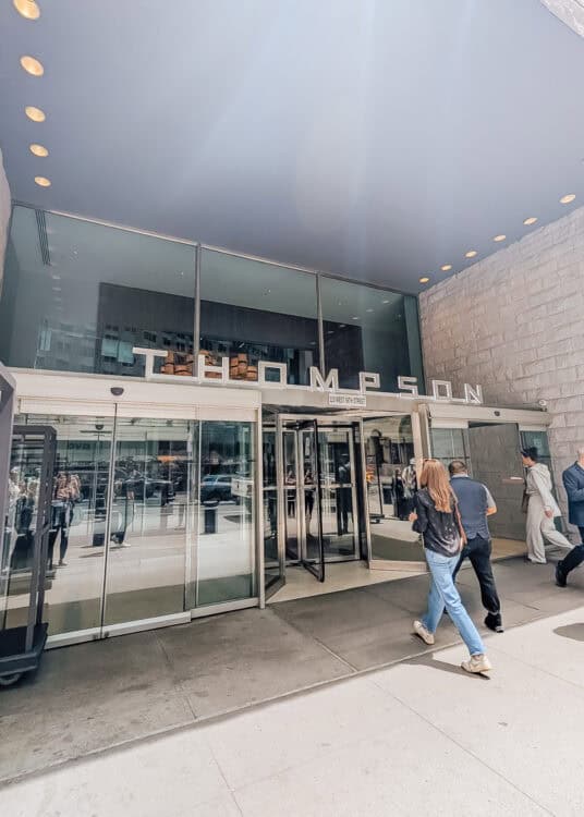 The entrance to Thompson Central Park New York features glass doors beneath a large metallic sign reading "THOMPSON" and "119 WEST 56TH STREET." Guests walk through the covered entrance of the Manhattan hotel with city reflections visible across the glass.