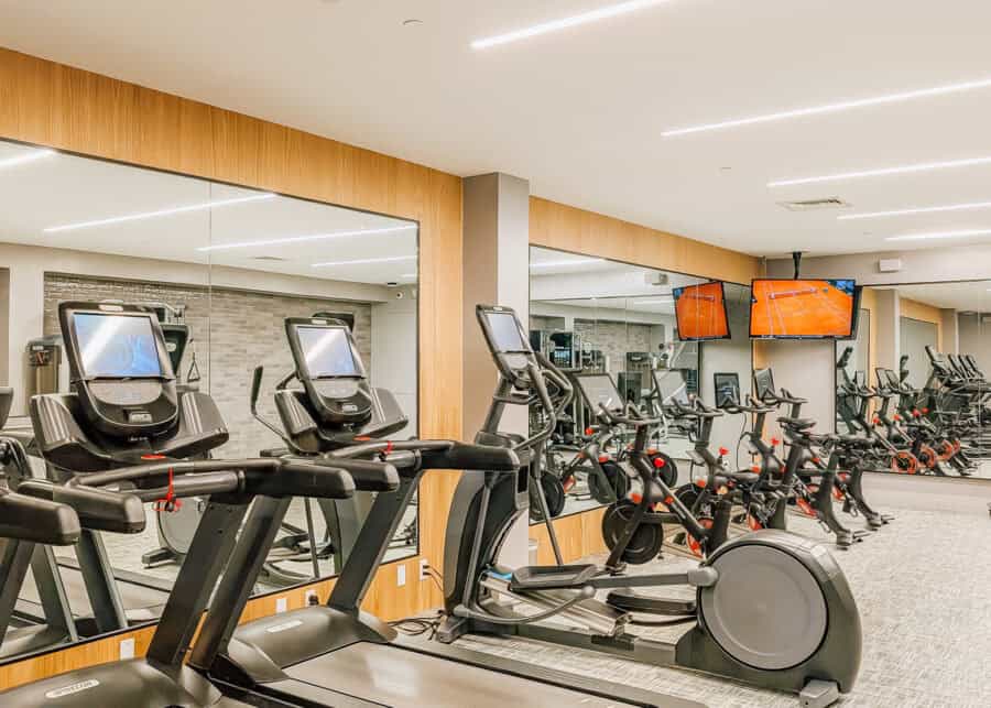 Fitness center at Thompson Central Park New York with treadmills elliptical machines and rows of stationary bikes. Floor to ceiling mirrors make the bright modern gym feel spacious while wall mounted televisions face the cardio equipment.