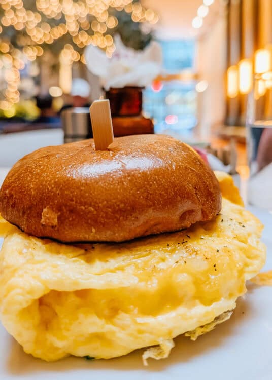 Breakfast sandwich at Thompson Central Park New York with a thick folded egg and melted cheese on a glossy toasted bun. Warm string lights glow softly in the blurred restaurant background.