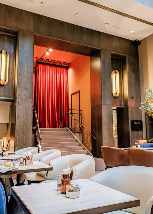 Restaurant at Thompson Central Park New York with cream and rust colored seating surrounded by dark wood walls and warm brass lighting. A staircase framed by dramatic red curtains rises behind the dining tables.