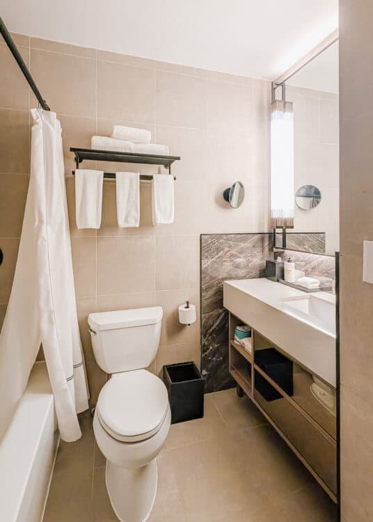 Bathroom at Thompson Central Park New York with a bathtub and shower curtain beside a toilet and long white vanity. Beige tile walls and a marble patterned backsplash are paired with neatly folded white towels and a large illuminated mirror.