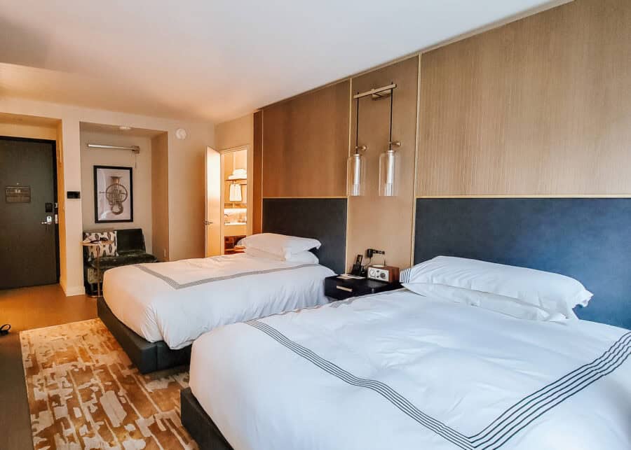 A guest room at Thompson Central Park New York features two beds with crisp white linens and dark blue upholstered headboards beneath modern hanging lights. A bedside table separates the beds while an open doorway reveals the brightly lit bathroom.