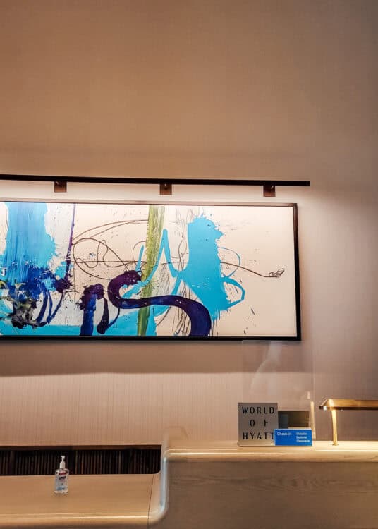 The check in desk at Thompson Central Park New York sits beneath a large abstract painting with bold blue green and purple brushstrokes. A counter sign reads "WORLD OF HYATT" beside a digital display reading "Check in Globalist Explorist Discoverist" and a brass desk lamp.