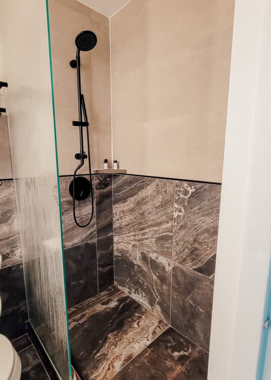 Walk in shower at Thompson Central Park New York with dramatic brown and cream marble tile and a glass partition. A matte black rainfall style handheld shower fixture contrasts with the neutral stone walls.