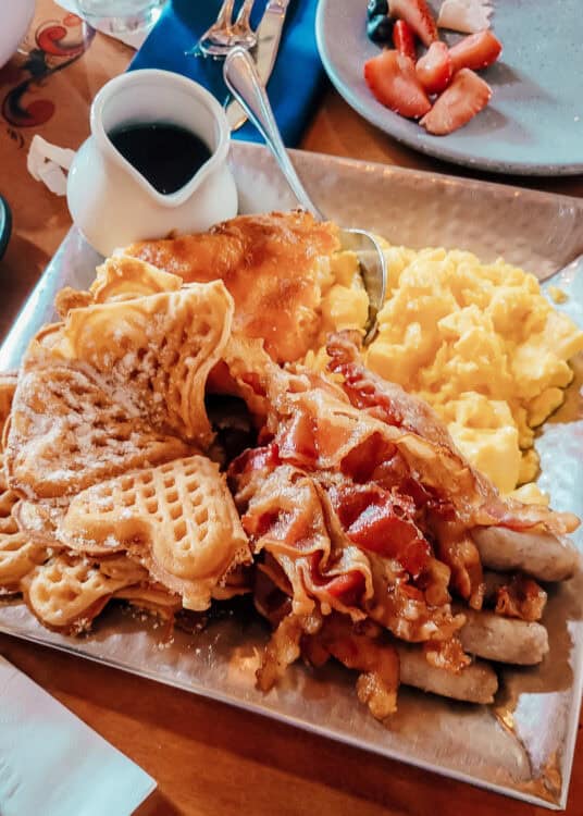 Large breakfast platter with waffles dusted in powdered sugar, crispy bacon, sausage links, scrambled eggs, and golden potatoes served with syrup on the side. Fresh strawberries and blueberries sit on a plate in the background beside glasses and silverware on the restaurant table.