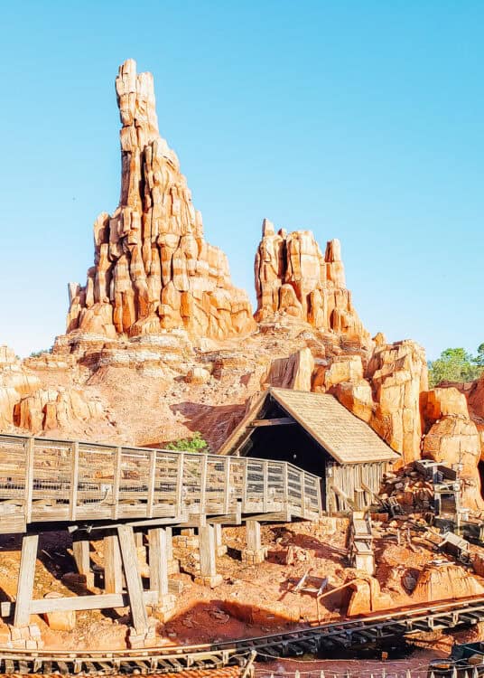 owering red rock formations rise above a wooden roller coaster track and mining structures in a desert themed attraction under a clear blue sky. Weathered wood bridges and rugged canyon scenery create the look of an old western mining town.
