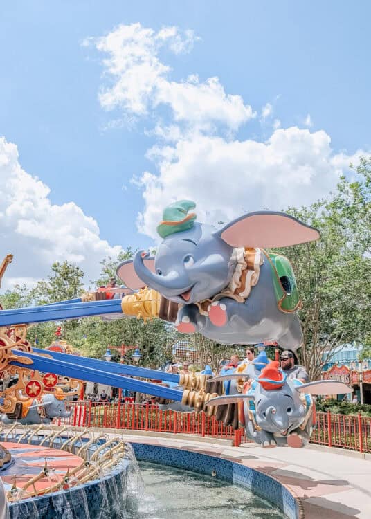 Dumbo themed flying ride with smiling elephant cars soaring above a fountain on a sunny day at Magic Kingdom. Families ride in colorful elephant vehicles while fluffy white clouds fill the bright blue sky overhead.